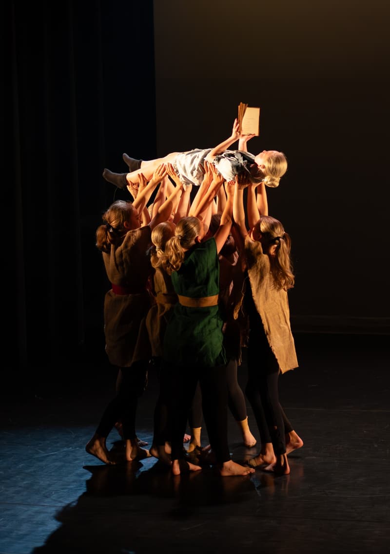 En gruppe dansere i kostymer av sekkestrie løfter en danser høyt over seg, hun holder en bok i varmt scenelys